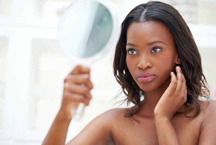 Black woman examining her face in a handheld mirror.