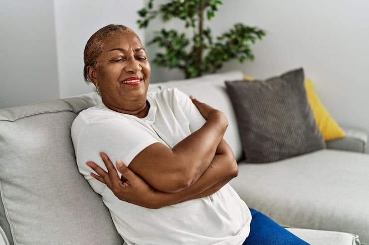 Smiling older woman hugging herself on a couch, showing self-love.
